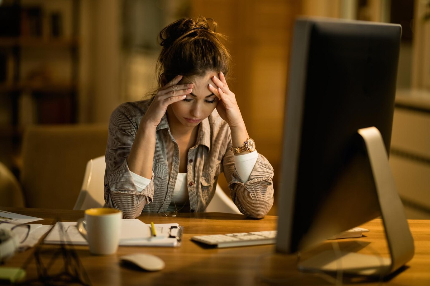 Uma pessoa de aparência cansada ou estressada em seu local de trabalho, com a cabeça apoiada nas mãos ou olhando para a tela do computador.
